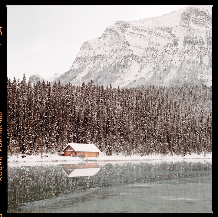 A wooden cabin sits by a partially frozen lake, surrounded by snow-covered pine trees and mountains.