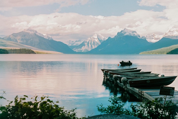 Several boats are docked on a calm lake with mountains in the background under a cloudy sky.
