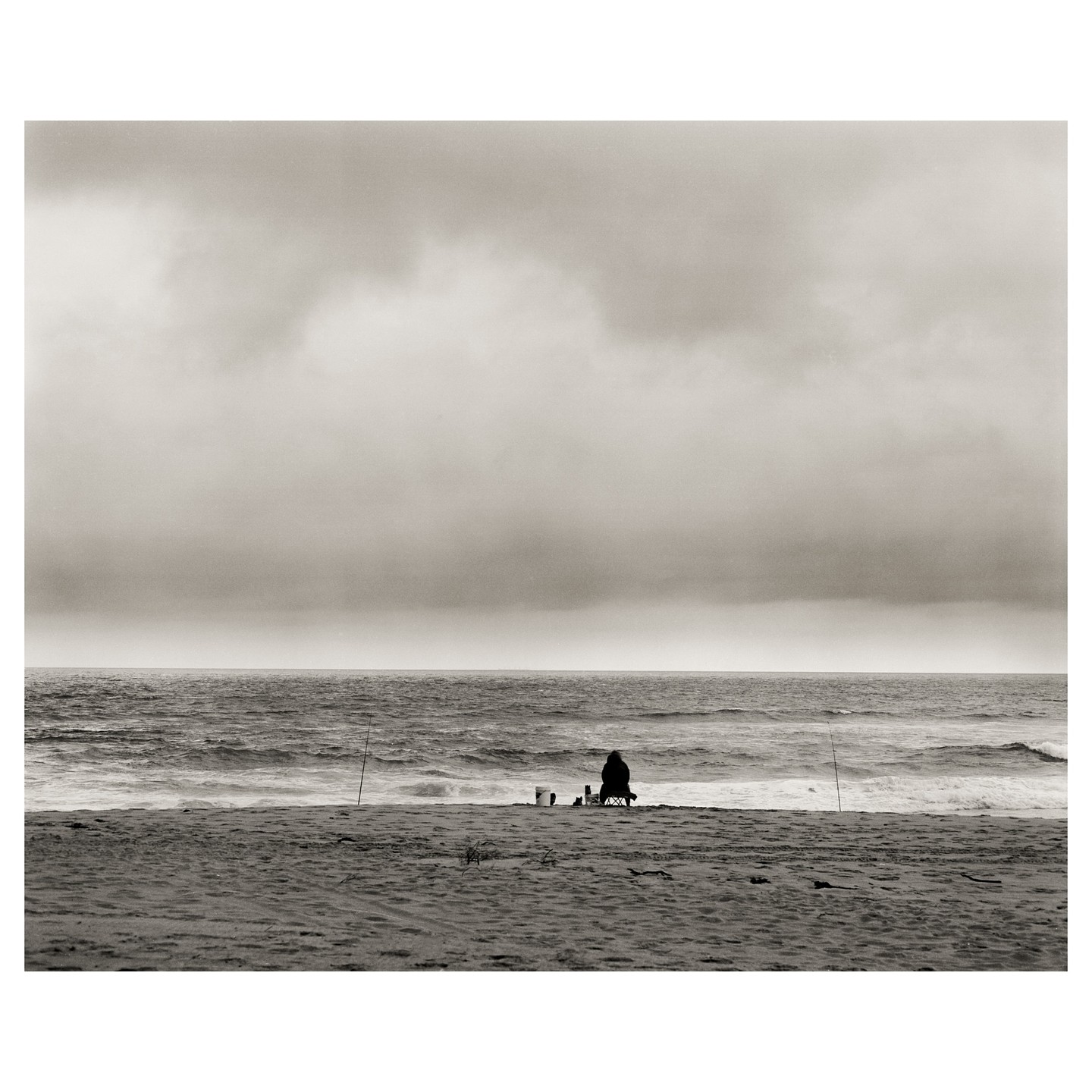 A person sits on a chair on a sandy beach, fishing in the ocean under a cloudy sky.
