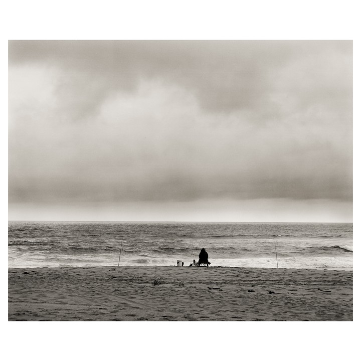 A person sits on a chair on a sandy beach, fishing in the ocean under a cloudy sky.