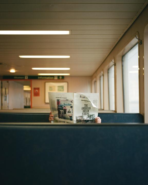 A person is reading a newspaper inside a vehicle with windows along one side.