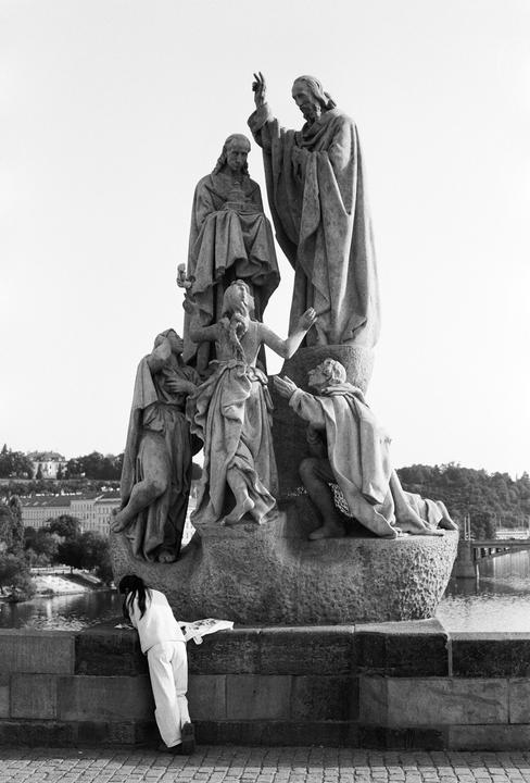 A person leans against a stone wall beneath a large religious sculpture on a bridge overlooking a river and city.