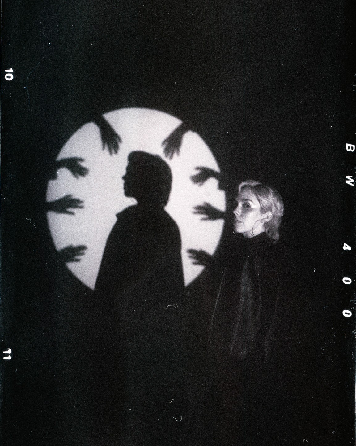 A woman stands next to a silhouette of a person with multiple hand shadows around a circular light.