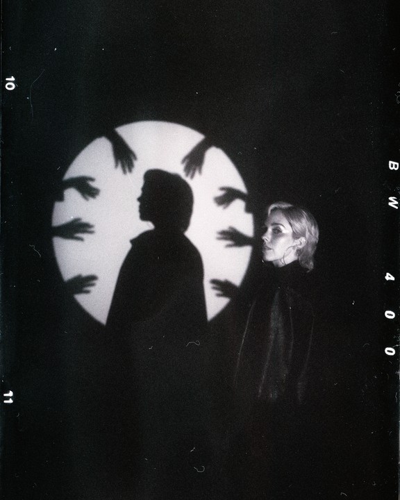 A woman stands next to a silhouette of a person with multiple hand shadows around a circular light.