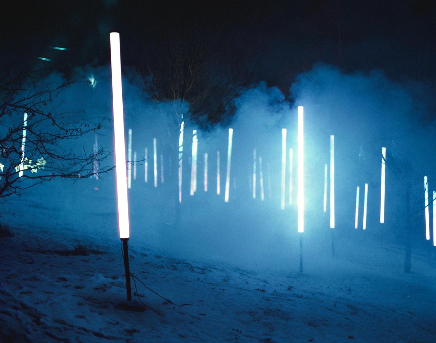 A snowy landscape at night is illuminated by many glowing light tubes and blue fog.