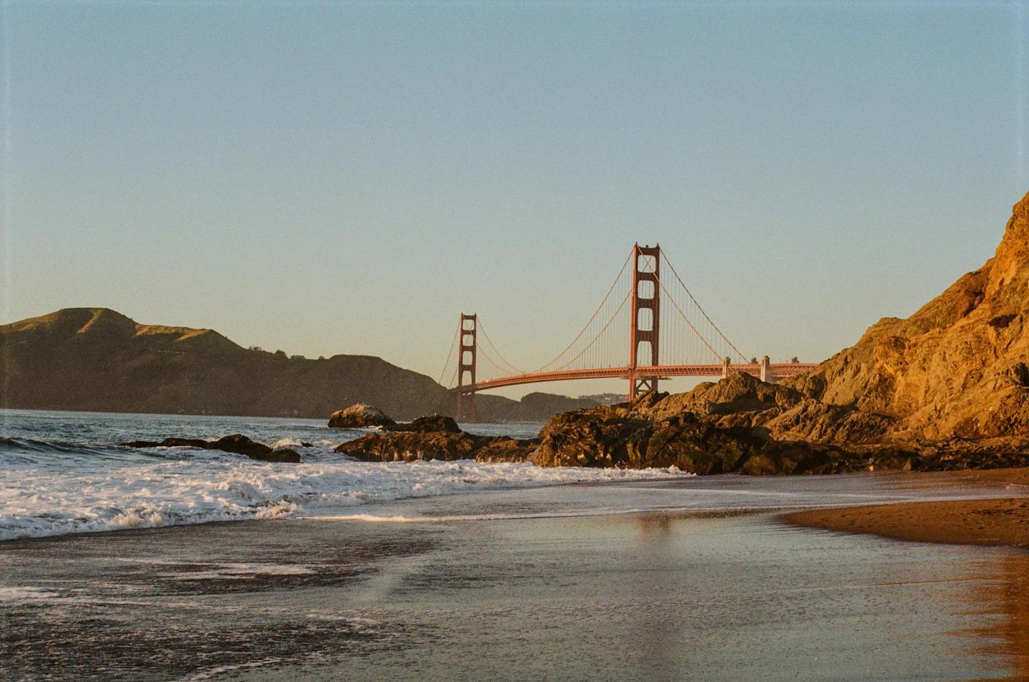 The Golden Gate Bridge is visible in the distance from a rocky beach with waves crashing on the shore.