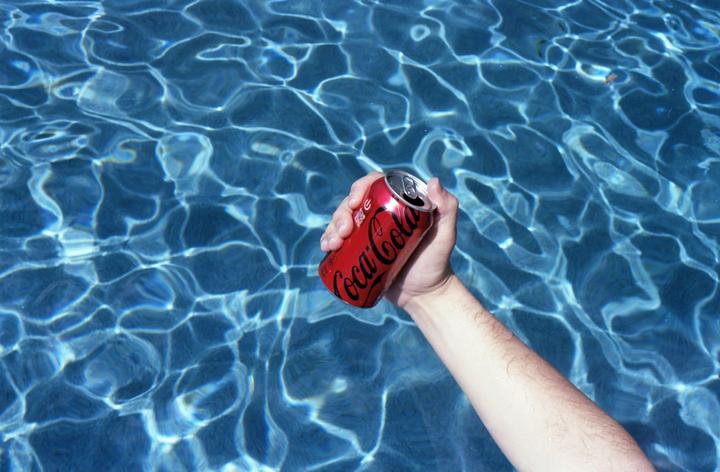 A hand holds a red Coca-Cola can above blue water with light reflections.
