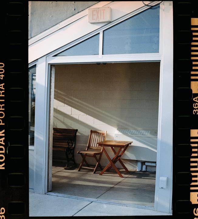 A wooden chair and table sit on a patio with a white brick wall and an exit sign above a window.