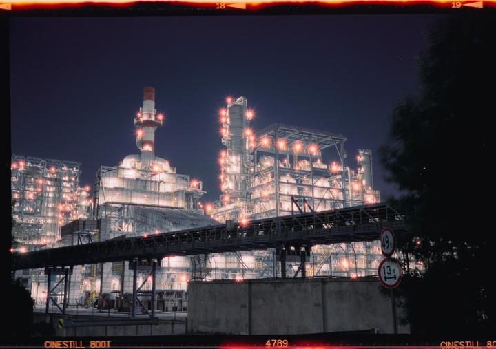 A large industrial complex with many lights is visible at night, with a dark tree in the foreground.