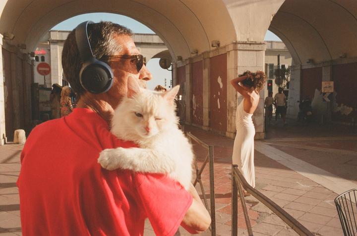 A man wearing headphones and sunglasses holds a white cat on his shoulder while a woman in a white dress stands under an archway.