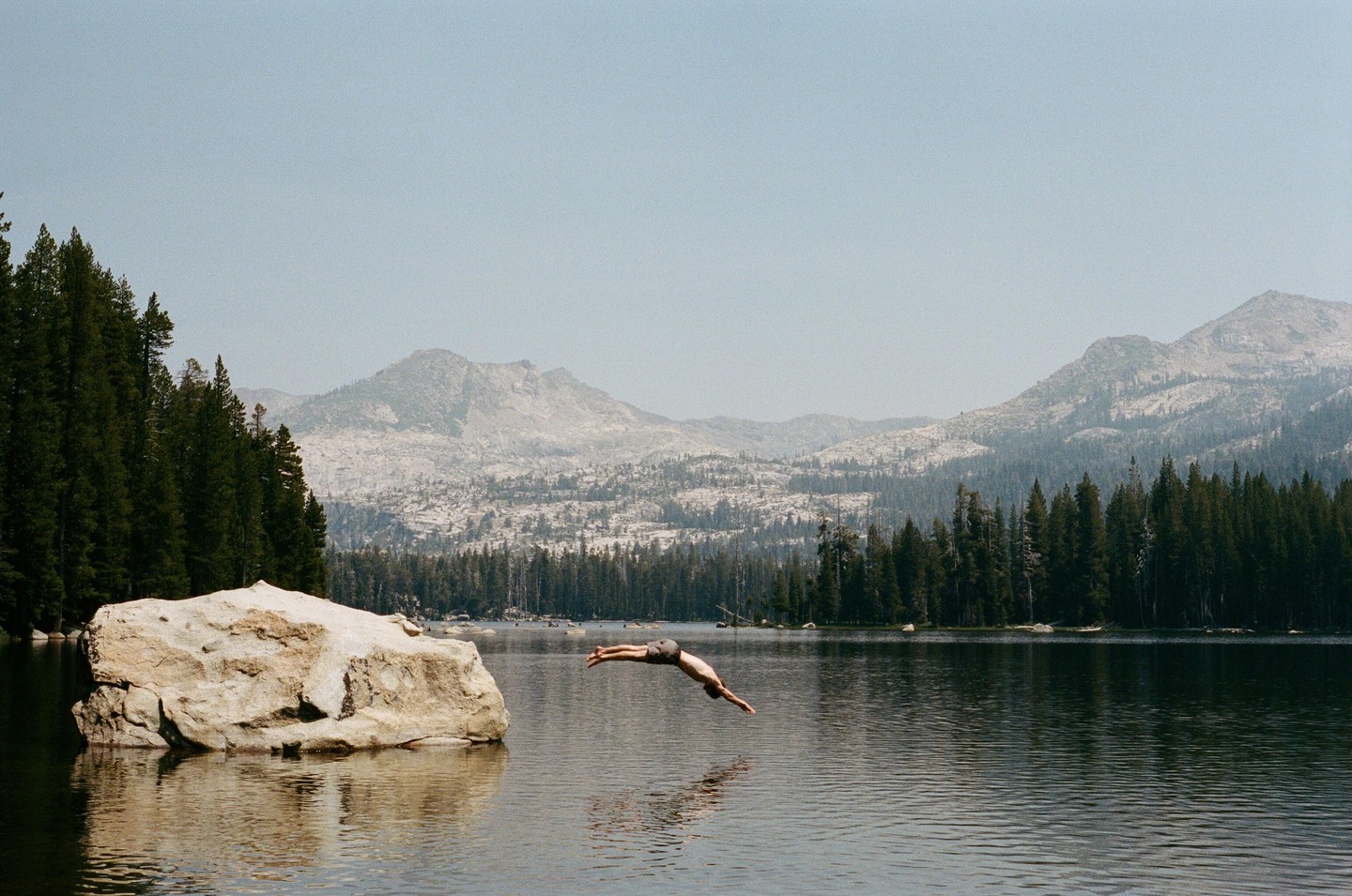 A man dives into a lake surrounded by a forest and mountains.