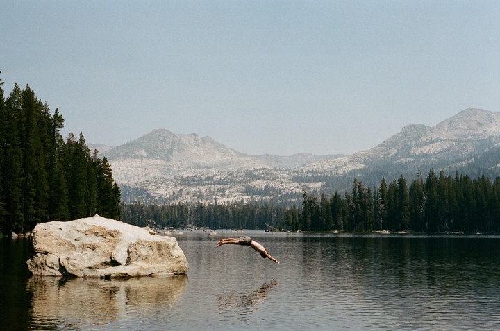 A man dives into a lake surrounded by a forest and mountains.