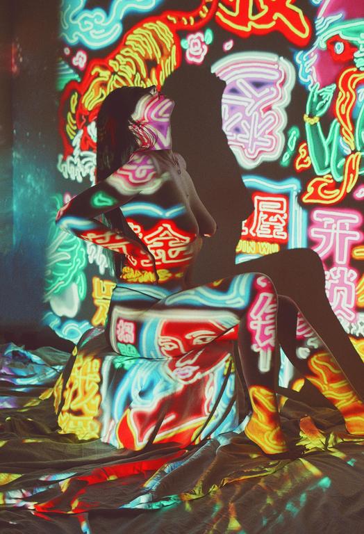 A woman sits with colorful neon light patterns projected onto her body and the surrounding surfaces.