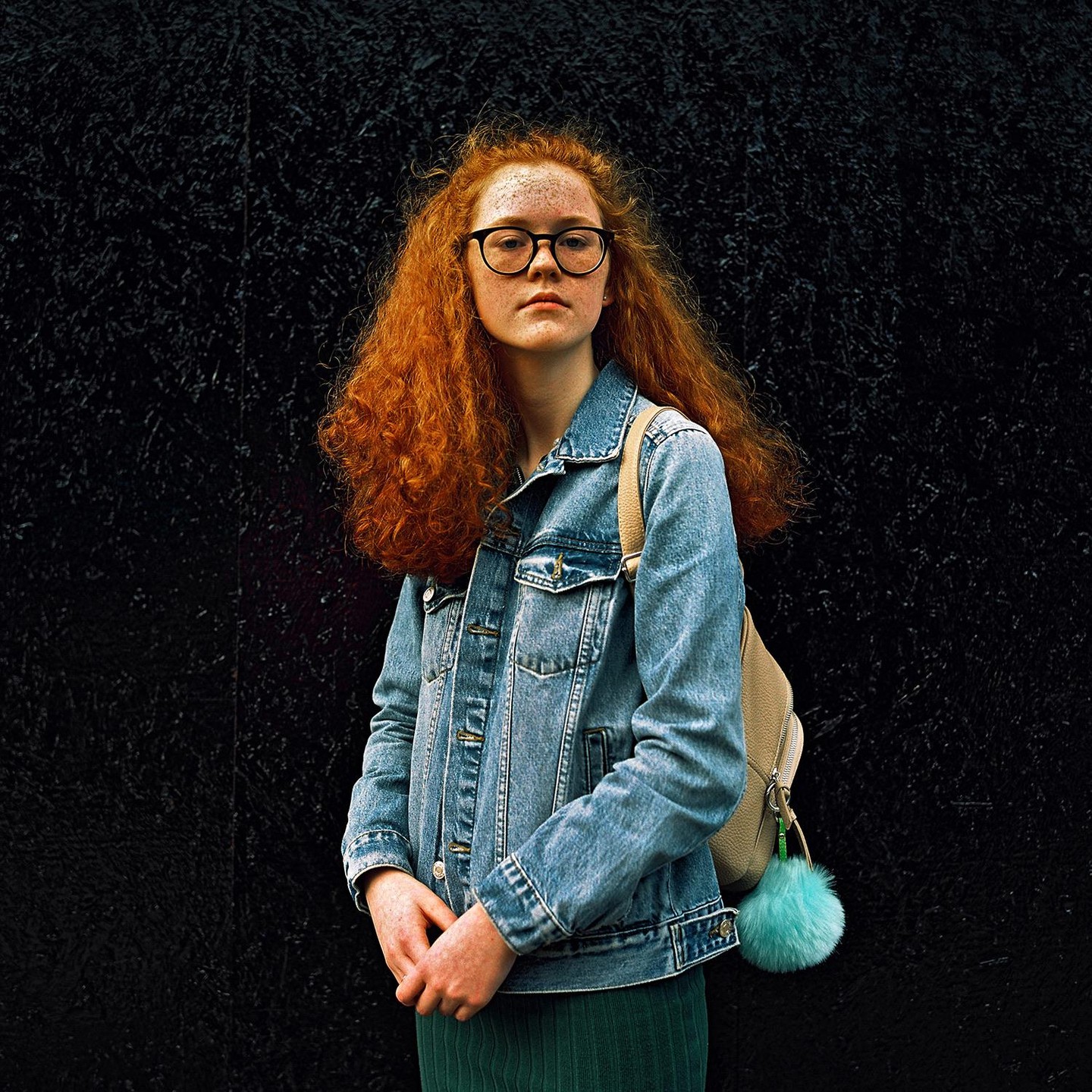 A young woman with long red curly hair and glasses stands in front of a dark textured wall.