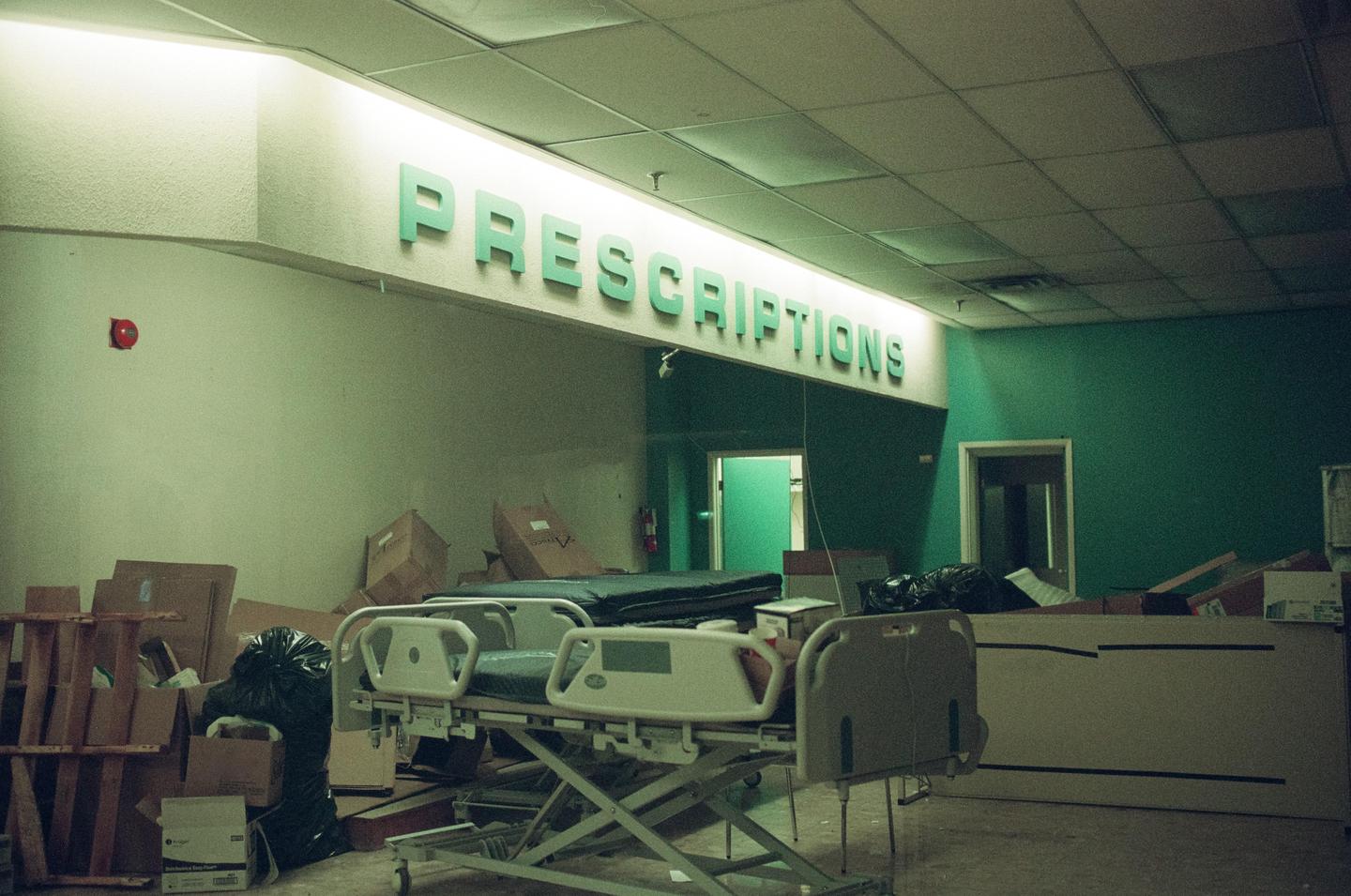 A hospital bed sits among cardboard boxes and trash bags under a sign that reads "PRESCRIPTIONS."