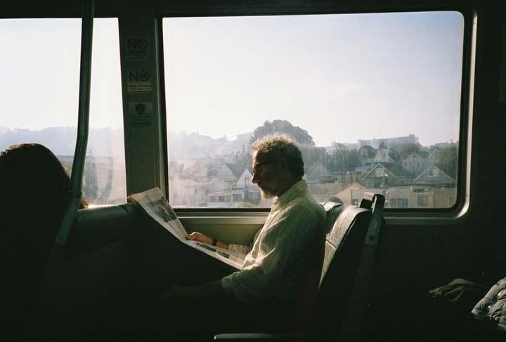 A man reads a newspaper on a train with a view of houses and hills outside the window.