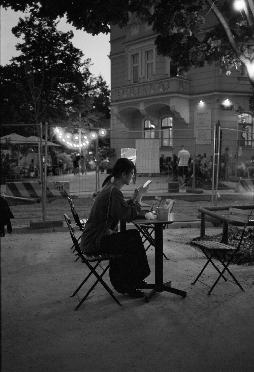 A woman sits at a table outdoors, looking at her phone, with a building and string lights in the background.