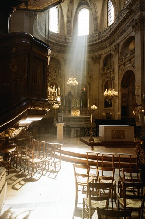 Sunlight streams through a high window into a large, ornate church interior with rows of wooden chairs and a central altar.