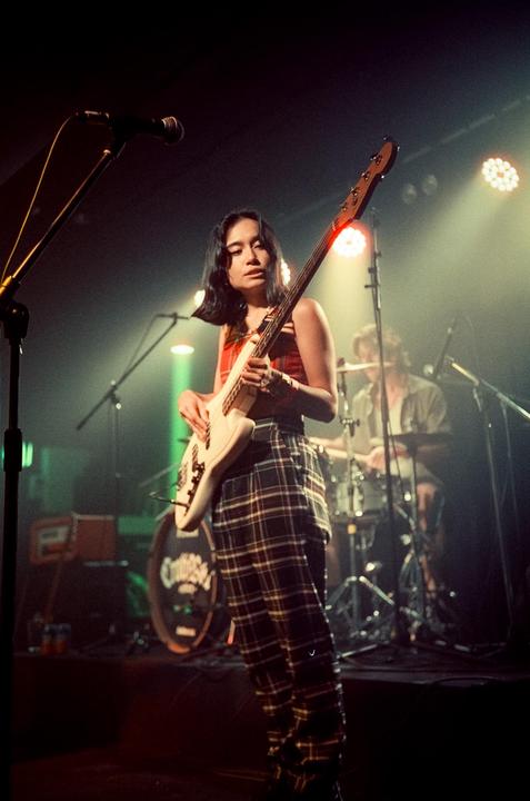 A woman plays a bass guitar on a stage with a drummer and microphone in the background.