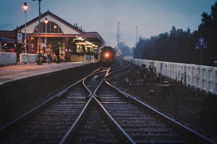 A train approaches a station at dusk, with tracks converging in the foreground.