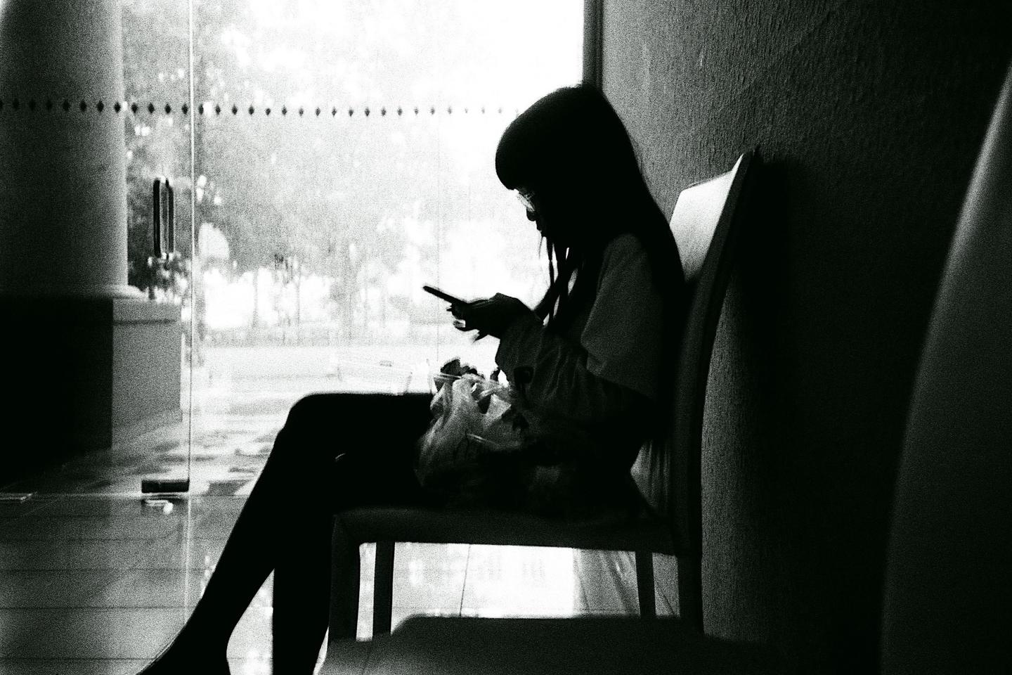 A woman sits in profile on a chair, looking at her phone, with a bright window behind her.