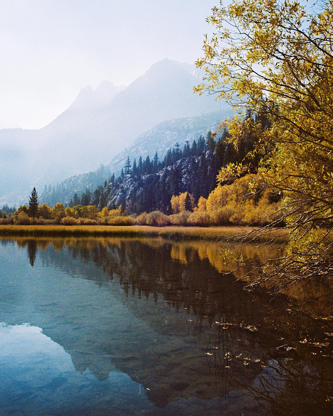 A lake reflects mountains and autumn trees under a hazy sky.