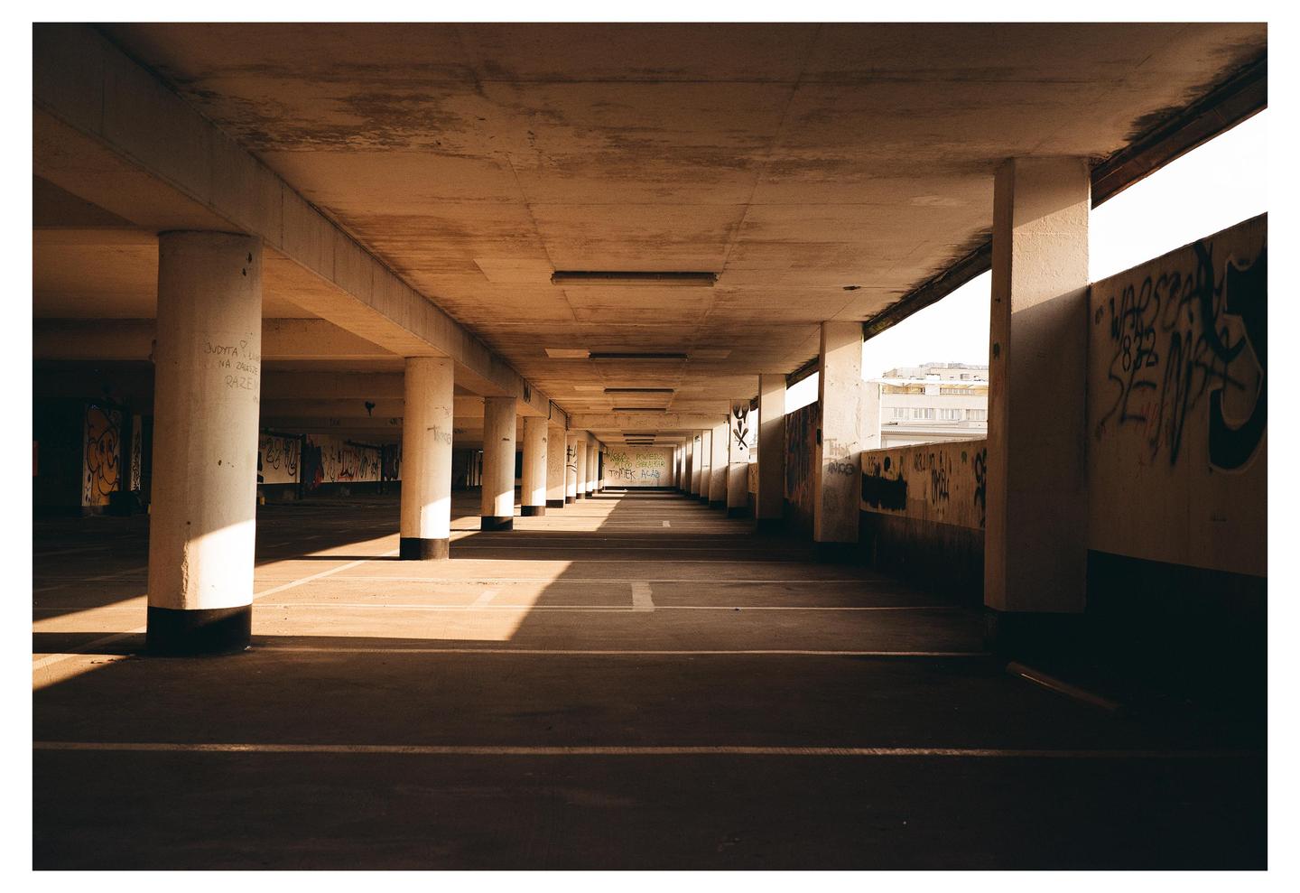 A long, empty parking garage with concrete pillars and graffiti on the walls is illuminated by sunlight creating strong shadows.