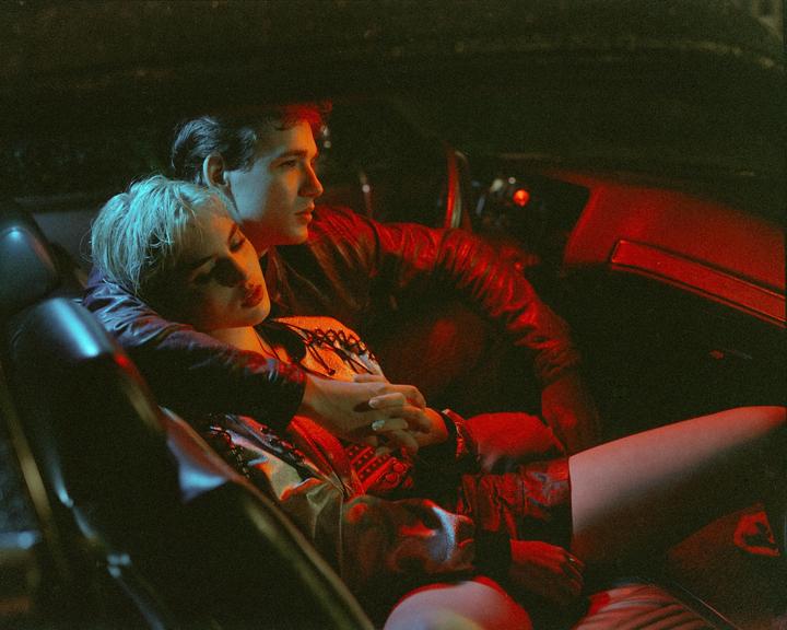 A man and a woman are sitting in a car at night, illuminated by red and blue light.