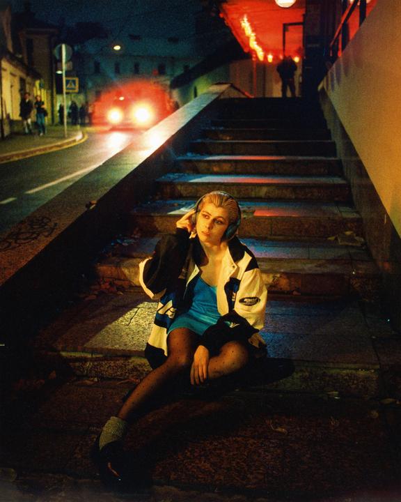 A woman wearing headphones sits on concrete steps at night with a street and buildings in the background.