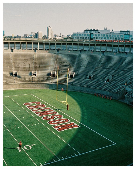 A football player stands on a green field with "CRIMSON" written in red letters, surrounded by empty stadium seating and a city skyline in the background.