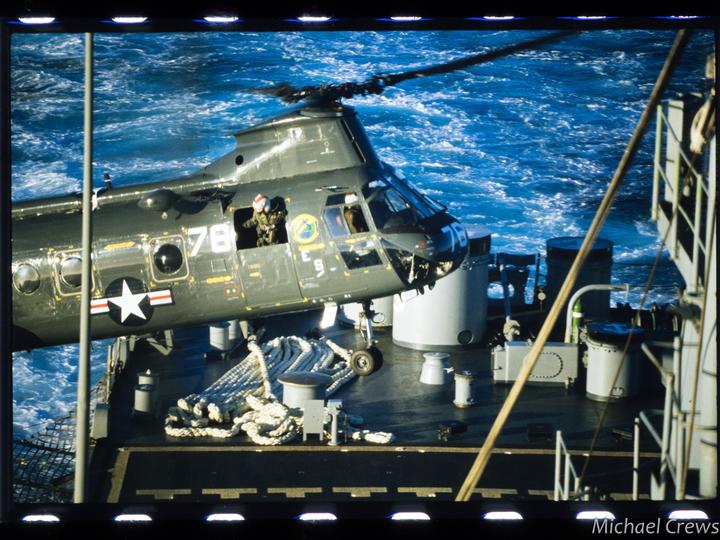 A military helicopter is shown on the deck of a ship with the ocean visible in the background.