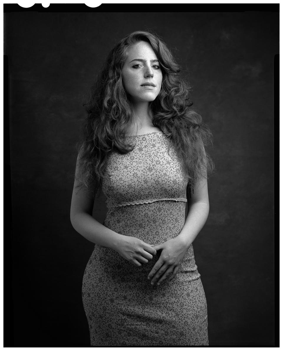A woman with long, curly hair and a patterned dress stands in a studio.