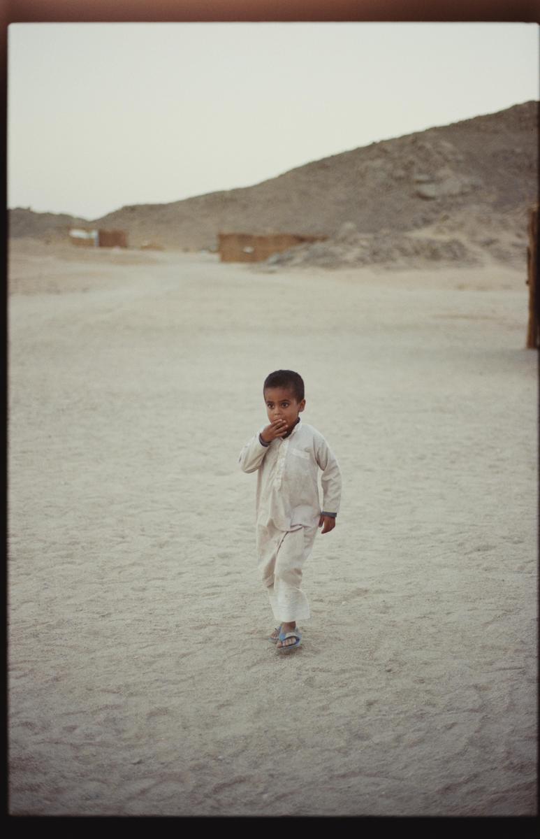 A child in a light-colored garment walks across a sandy desert landscape with hills and buildings in the background.