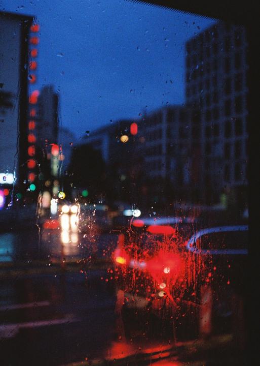 A rainy city street at night is viewed through a window covered in water droplets, with blurred car lights and buildings in the background.
