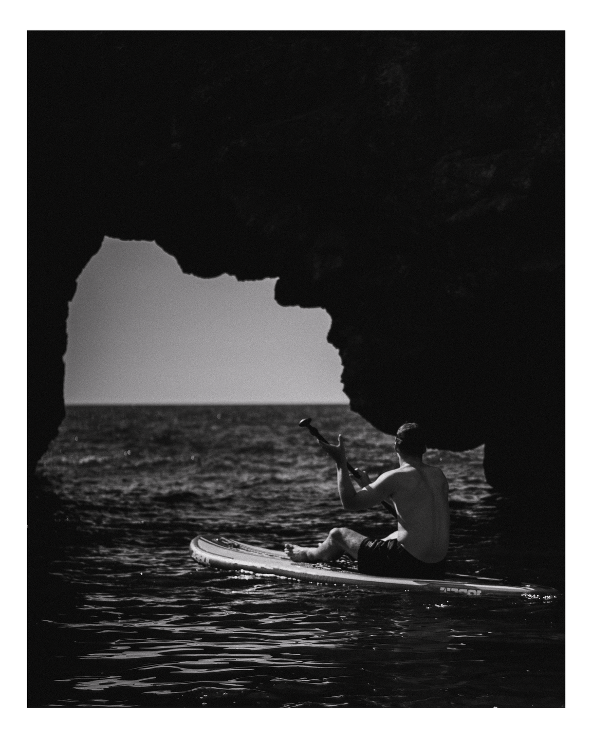 A man sits on a paddleboard in the ocean, framed by a dark rock archway.