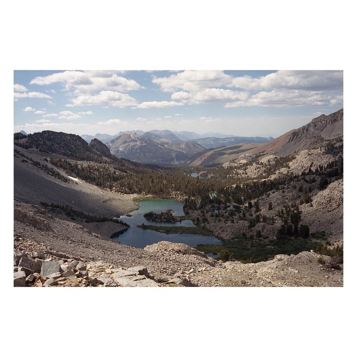 A high-angle landscape view shows a valley with several lakes and evergreen trees surrounded by mountains under a cloudy sky.