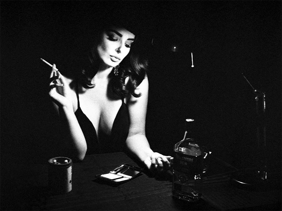 A woman in a dark dress holds a cigarette while sitting at a table with a bottle of whiskey and a compact.