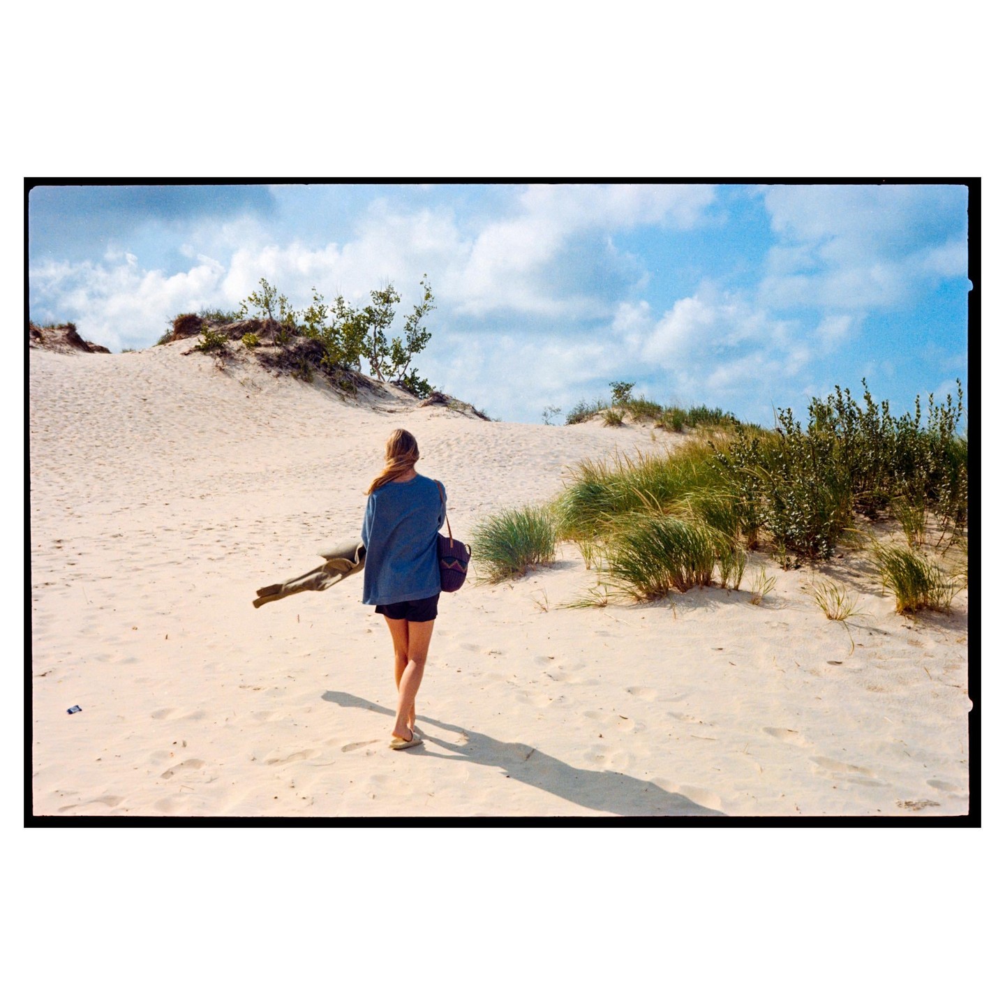 A woman walks away from the camera across sand dunes with sparse vegetation under a blue sky with clouds.