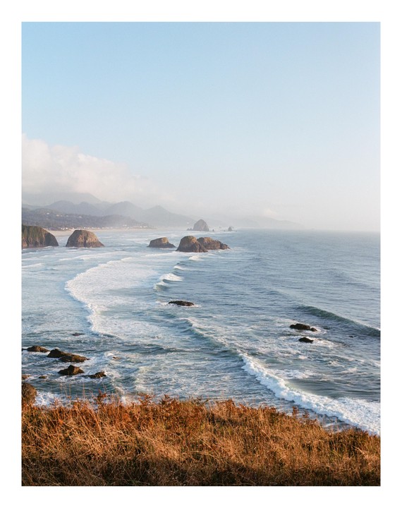 Waves crash on a rocky coastline with a hazy sky and mountains in the distance.