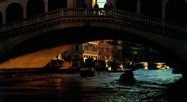A bridge with people on top casts a shadow over a canal with boats and buildings in the background at sunset.