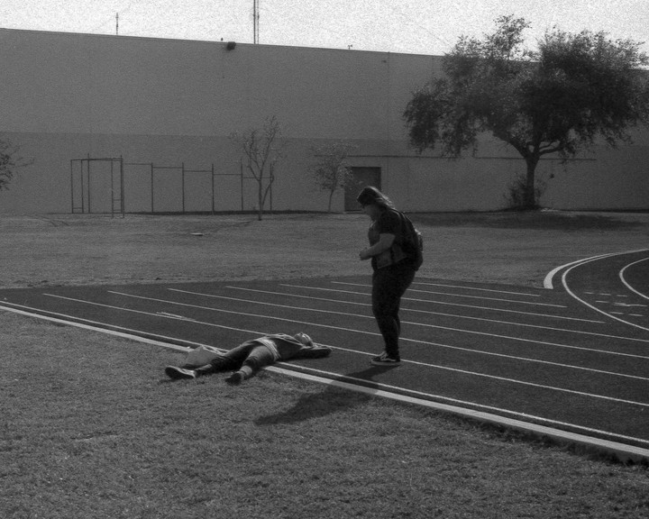 A woman stands on a running track while another person lies on the grass next to it, with a building and trees in the background.