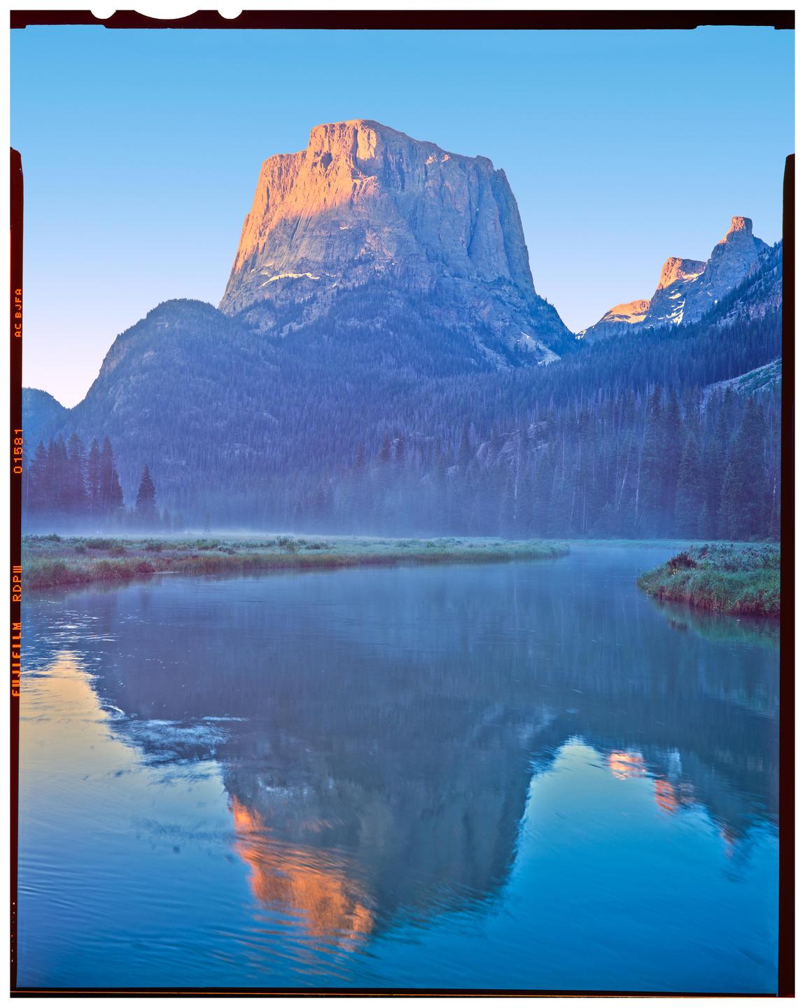 A large mountain with a sunlit peak is reflected in a calm body of water, with mist rising from the water's surface.