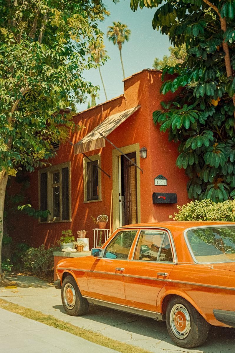 An orange car is parked in front of an orange house with green foliage and palm trees.