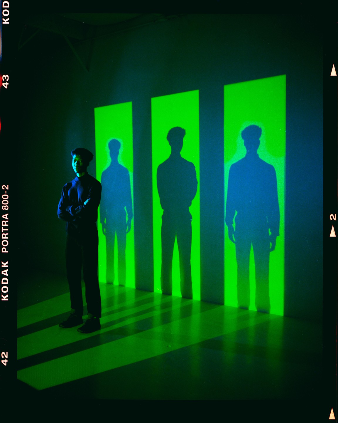 A man stands in a dark room with three green rectangles projected on the wall behind him, each containing a shadow of a man.