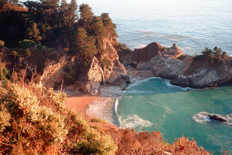 A waterfall cascades onto a sandy beach next to a turquoise ocean, surrounded by cliffs and pine trees.