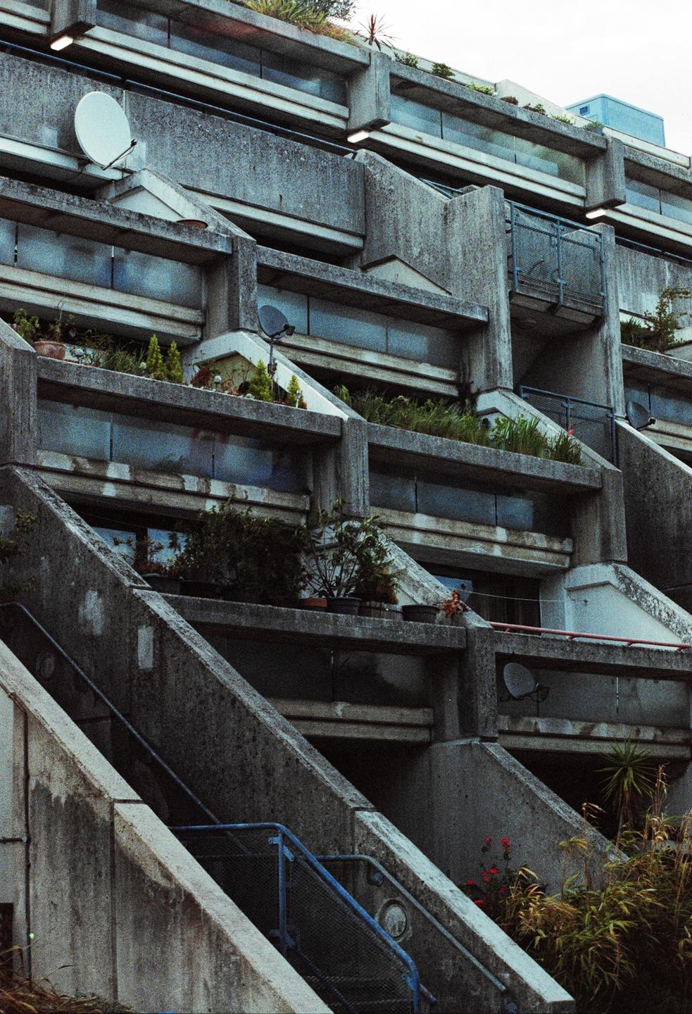 A brutalist concrete building with plants on balconies and satellite dishes on its facade is shown from a low angle.
