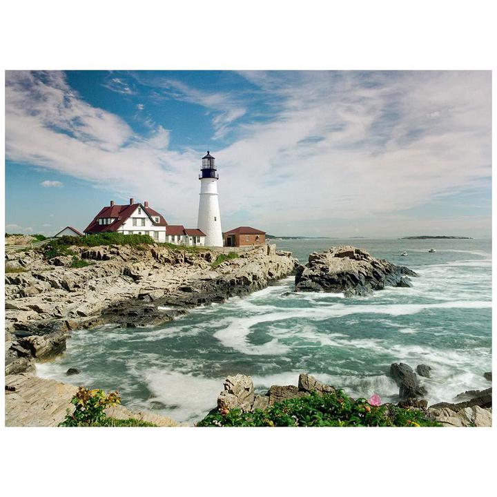 A white lighthouse stands on a rocky coast next to a large white building with a red roof, with the ocean and a cloudy sky in the background.