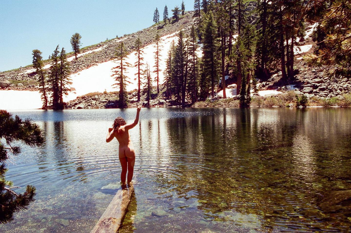 A nude woman stands on a log in a clear lake with snow-covered mountains and pine trees in the background.