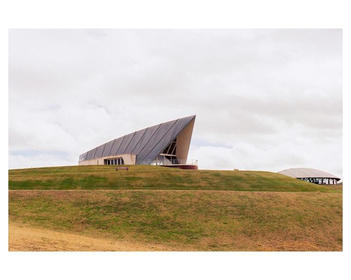 A modern building with a sharply angled roof sits atop a grassy hill under a cloudy sky.