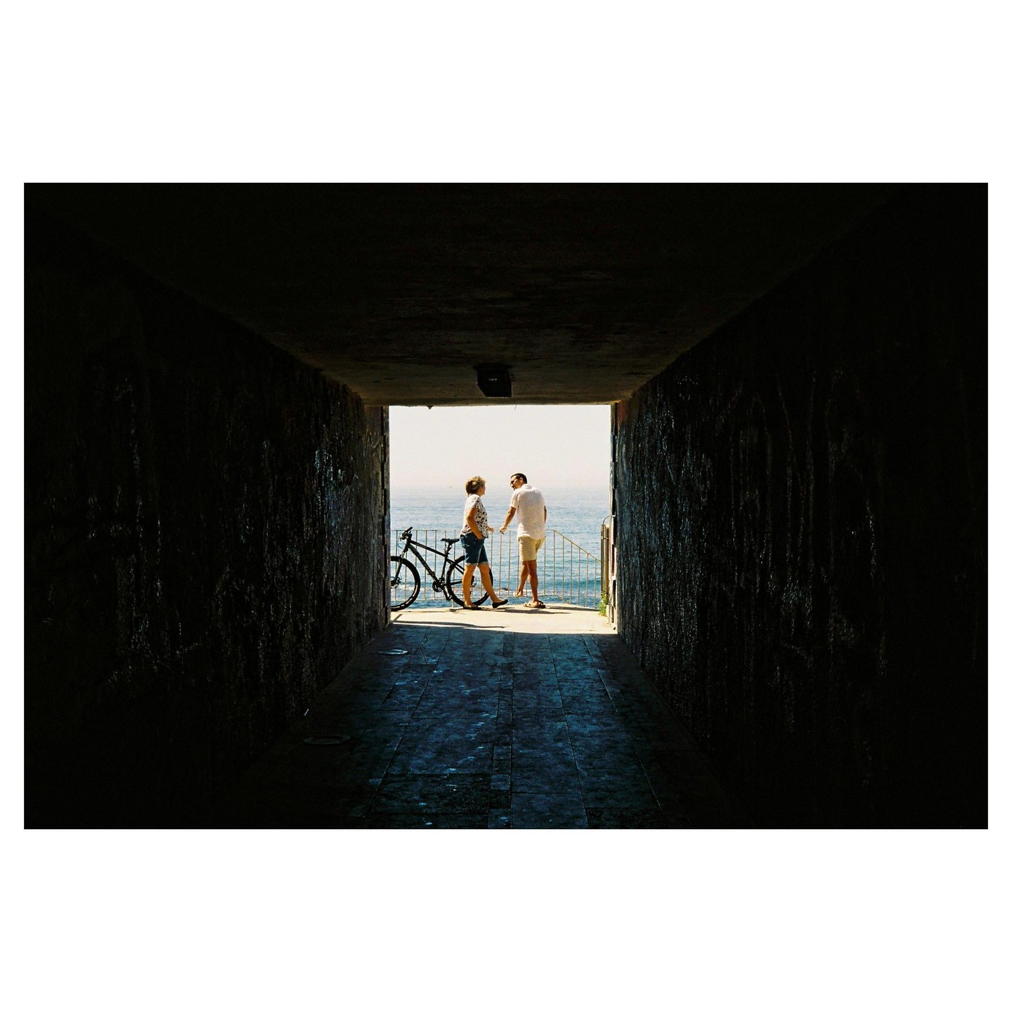 A couple stands with a bicycle at the end of a dark tunnel, looking out at the ocean.